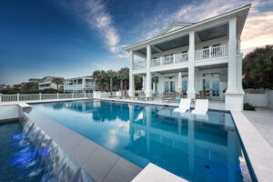 Luxury residential swimming pool with a modern waterfall edge and built-in lounge chairs, located in front of a large coastal-style home in Southwest Florida.